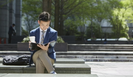 広場の椅子に座り手帳を読むビジネスパーソン
